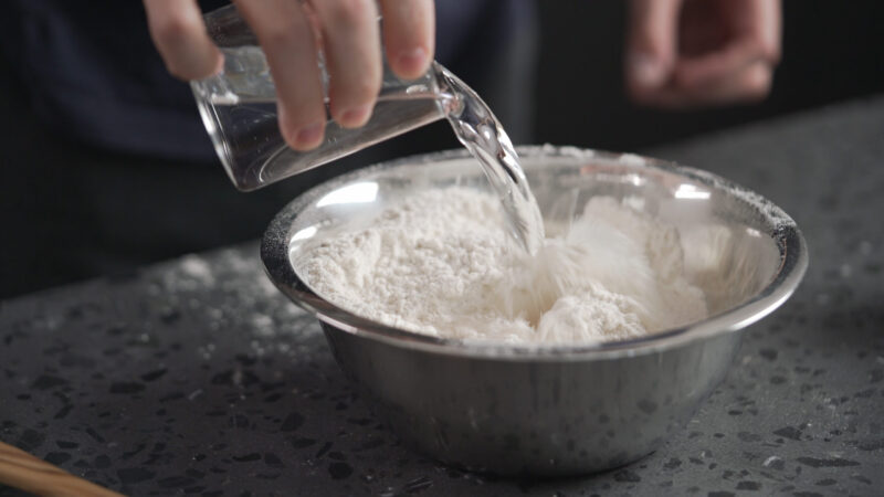 man adds wet ingredients into flour in steel bowl on concrete countertop